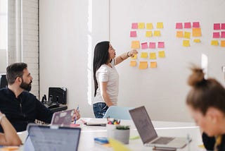 Sessão de workshop, onde há pessoas sentadas ao redor de uma mesa, com seus computadores e uma mulher, em pé, com post-its.