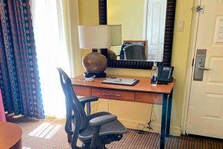 This photo is of a hotel room. There is a large wooden framed mirror and a desk with a chair and a lamp, laptop, and a bottle of water. The laptop is closed and the window curtain is partially open and the sun light is bright.