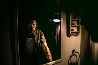 A fairly muscular guy is in a dark room with a single light. He looks angry and frustrated.
