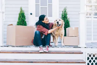 A woman pets a dog while sitting next to moving boxes.