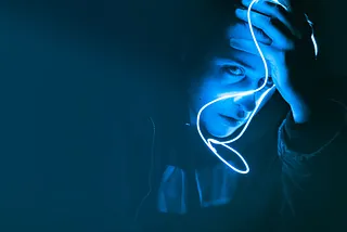 Person in a dark background with a cobalt blue light on their face, looking despondently at the camera.