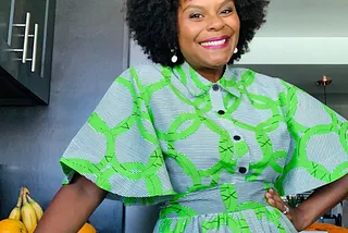 Tabitha Brown posing in a kitchen while wearing a bold graphic print dress.