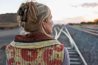 A woman facing away from the camera while standing on train tracks.