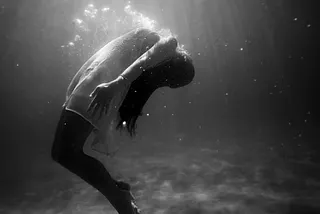 A black-and-white image of a person submerged underwater, arching backward in a weightless, dreamlike pose. Sunlight filters through the surface, casting ethereal beams and illuminating the surrounding stillness. The scene evokes a sense of struggle, surrender, and quiet introspection.