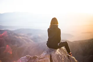 Woman sitting on the top of a mountain overlooking a body of water with islands.
