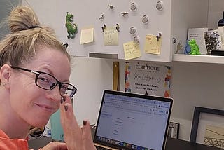 Mia sits smiling in her favorite orange sweatshirt at her laptop, working on her writing on her laptop. She is in her office, with lots of decorations around and a copy of her book, Becoming a Personal Trainer, on her desk.