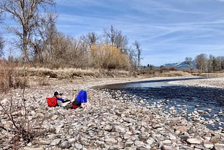 The author reading a book on a pebble beach next to a channel of the Clark Fork River in Missoula, MT