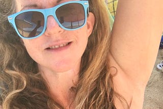 Selfie midshot of the female story author grinning at the camera, wearing baby blue sunglasses and a black bikini top, while seated in the shade