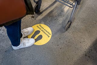 A photo of a person standing on a sticker that says “practice social distancing” at a grocery store.