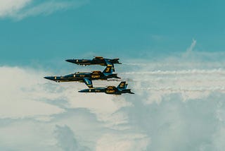 A group of three blue and yellow US navy fighter jets flying through a cloudy sky.