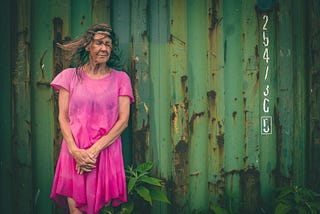 Photograph of an older woman wearing a pink dress and standing in front of a rusted shipping container