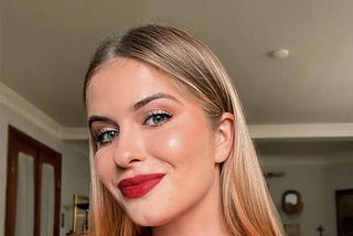 A selfie of Katie wearing a red one-shoulder dress and a gold necklace. She has blonde hair, blue eyes, bright red lipstick, and appear to be in a home setting.