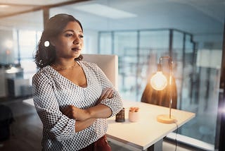 A woman stands in her office alone at night with her arms crossed, thinking about her job.