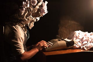 Quirky photo symbolizing writer’s block. Person sitting at typewriter with a bunch of crumpled paper.