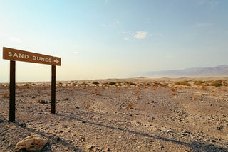 An image from Unsplash showing a desert landscape with a single, somewhat forlorn road sign saying “Sand Dunes” with an arrow pointing toward the horizon.