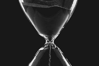 This is a black and white photograph of a glass hourglass with the sand running down set against a black background.