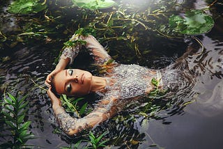 Beautiful woman is immersed in water on her back in seaweed. She is face up, eyes closed. Beautiful makeup, elbows bent, hands resting on the top of her head. Magic, beauty, fantasy aesthetic. Captivating.