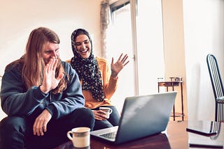 A couple smiles and waves at the camera on their laptop.