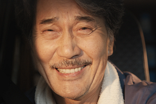 A close-up of Hirayama smiling and half-crying. He has a large almost pained grin, his lips are pursed and you can see his teeth. He has a small moustache. You can see the car headrest behind his head. The sun is shinning into his face. You can only see the top part of his blue overalls. His overalls have a white collar.