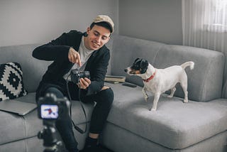 A young man sets up his camera and tripod on his couch. His dog is on the couch looking at him hold his camera.
