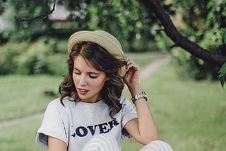 Image of a girl with long hair sitting on a drystone wall with trees and a garden in the background. She wears a hat, a grey t-shirt with logo ‘Lover’, grey and white striped trousers and white tennis shoes