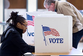 A photo of a black woman voting at a poll. There is a divider on the desk that reads “VOTE” with a U.S. flag.