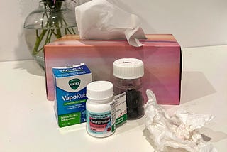 A collection of medicines, including Vicks VapoRub, a box of tissues and vase of mauve chrysanthamums.