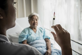 A photo of a nurse holding up a syringe with an elderly woman sitting in a hospital bed in the background.