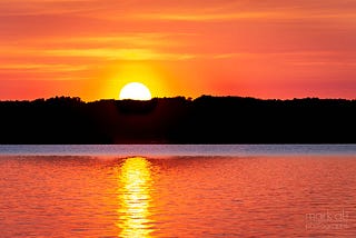 The sun rises over (and reflects off of) a calm lake in upstate New York.