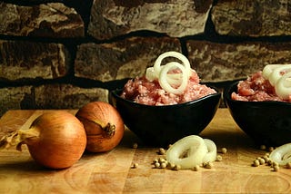 Two unpeeled onions beside two bowls of raw mincemeat with sliced onion rings on top.