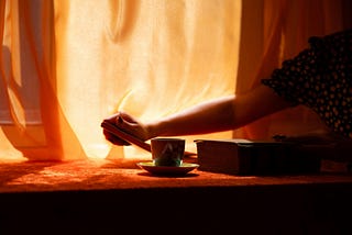 Amber light glows through sheer curtains, backlighting the table where a woman reaches for a cup sitting neatly in its saucer.
