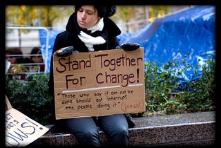 A woman in jeans and a black coat and black and white scarf sitting on a concrete bench at Occupy Wall Street, with a blue tarp in the background. She is holding a sign that reads “Stand together for change! Those say it can not be done should not interrupt the people doing it. Join us!”