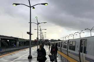 An open-air BART platform with a few passengers walking on a gray day.