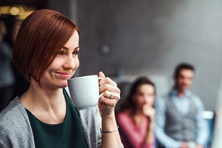 A woman smirks as she holds a coffee mug up to her face in front of 2 coworkers.