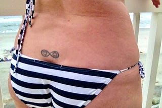Color photo of the author’s lower back and dark-blue with white striped string-bikini-clad buttocks, while showing her tattoo just above the middle of her bum-crack: a black ‘infinity’ with 2 black-outlined red hearts, one facing up and the other facing down.