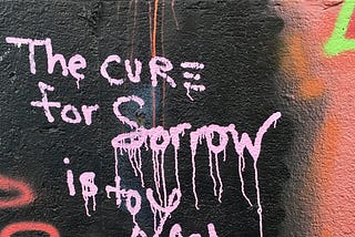 Light pink colored spray-painted graffiti writing on a wall of mainly black with some other colorings wall stating “The cure for sorrow is to learn something”.