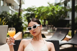 Confident young ethnic lady drinking juice lying on sunbed at poolside