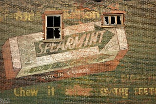 An old, faded ad for Wrigley’s spearmint gum painted on a brick wall pierced by two broken windows.
