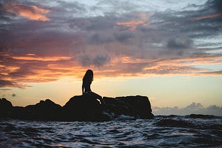 A young woman sitting alone on a rocky outcrop, surrounded by the  ocean. The sky is heavy with dark clouds lined in shades of orange and red by the setting sun.