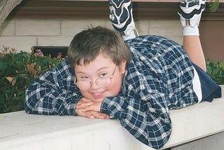 A young boy with Down syndrome laying on his tummy on a bench with his chin resting on his hands smiling at someone of to the side.
