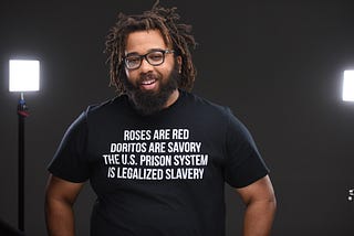 Kurt Evans in front of a black backround and studio lights, wearing a shirt saying the US prison system is legalized slavery.