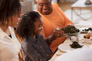 A smiling grandmother serves vegetables to her granddaughter at the dining table.