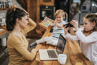 An exasperated mother trying to work at a laptop while her 2 children hold up an abacus and phone respectively.