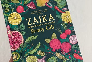 A hand holding the book “Zaika” by Romy Gill, which has illustrations of various vegetables and fruits on a dark green cover