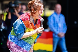 Kamala Harris speaking at an event on stage, wearing a sparkly button-up shirt with the rainbow pride flag on it