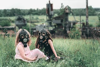 Two young girls sit on grass wearing gas masks.