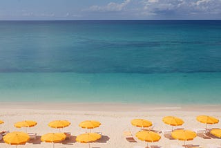 A beach with two rows of large yellow umbrellas, with a clear blue ocean in the background.