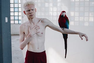 An arty photograph of a shirtless man with his outstretched arm, which has a red-and-blue parrot perched near the elbow.