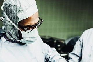 Two doctors operating on a patient. One doctor’s face is in frame and he is wearing scrubs, a mask, and glasses. Background wall is green. Medical equipment can be seen.
