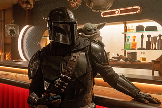 The Mandalorian is standing next to a bar wearing his full Mandlorian armour which is silver and sleek and reflects almost every light soruce. On his left shoulder is Grogu, he is wearing a brown robe. The Mandalorian has his left hand on the bar top. Behind the bar are shelves of random differently coloured bottles. The Mandalorian and Grogu are looking at something off screen to the right.